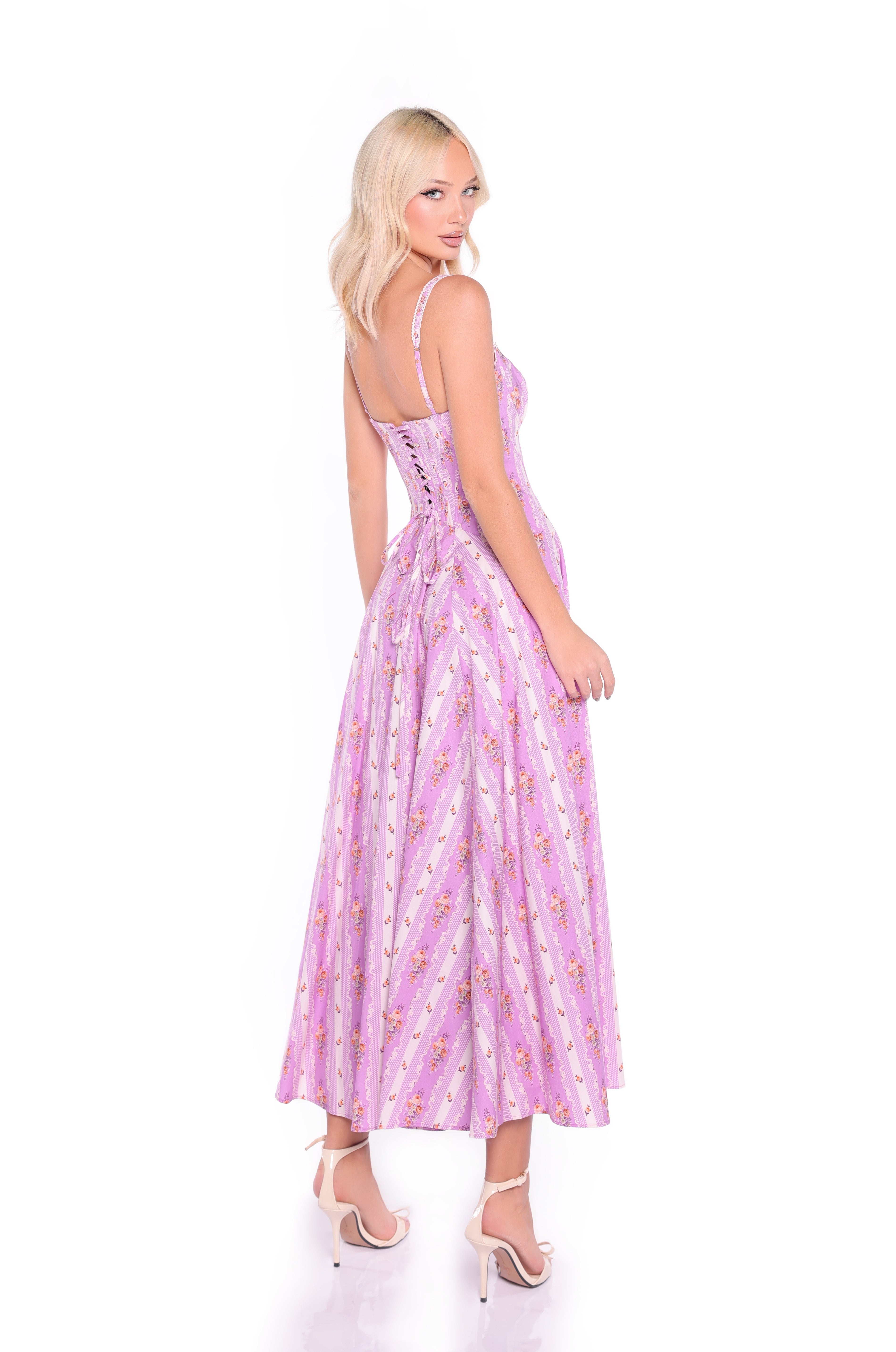 Woman in the Rosella Lavender Lilac Floral Casual Midi Dress with spaghetti straps, corset bodice, and lace-up back, standing in heels and looking over her shoulder against a white background.